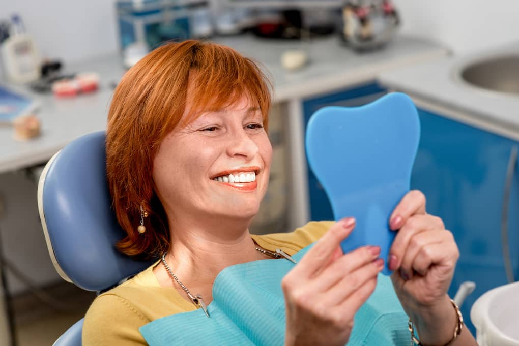 Smiling senior woman with new dental implants sitting in the dental office and looking at the mirror